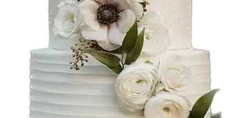 A three-tiered white wedding cake with textured frosting, decorated with white roses and a large anemone flower, displayed on a gold cake stand.