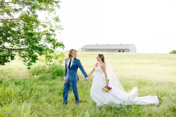 A bride and groom walking hand-in-hand through a grassy field with a white barn venue in the background.