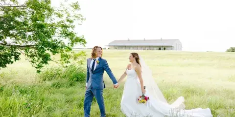 A bride and groom walking hand-in-hand through a grassy field with a white barn venue in the background.