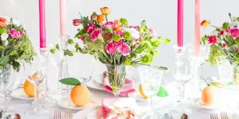 A professionally set event table featuring white plates, silver flatware, crystal glassware, pink napkins, and floral centerpieces.