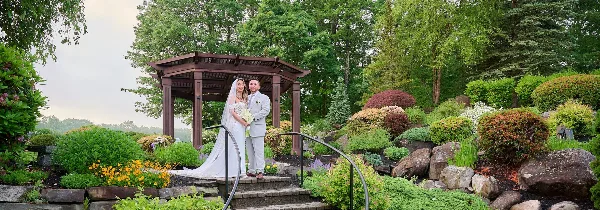 A bride and groom standing in a garden setting with a wooden gazebo and lush greenery.