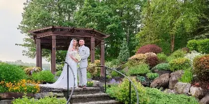 A bride and groom standing in a garden setting with a wooden gazebo and lush greenery.