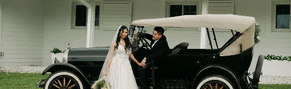 A bride and groom posing together next to a black vintage car in front of a white building.