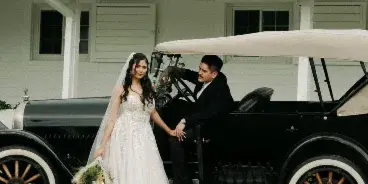 A bride and groom posing together next to a black vintage car in front of a white building.