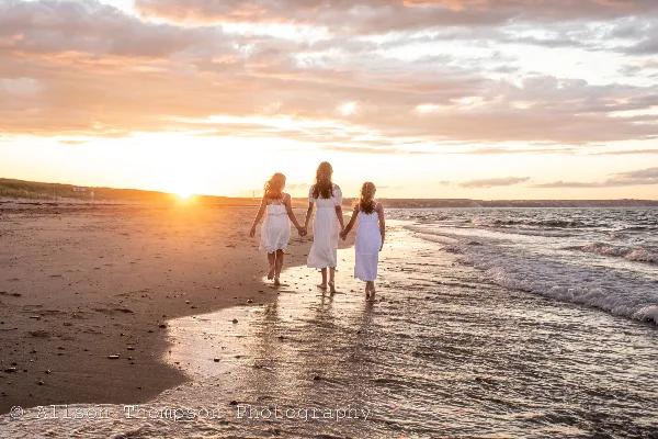 Three women in white dresses walking hand-in-hand on a sandy beach at sunset.