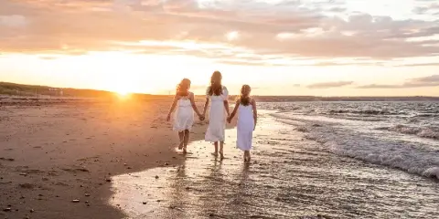 Three women in white dresses walking hand-in-hand on a sandy beach at sunset.
