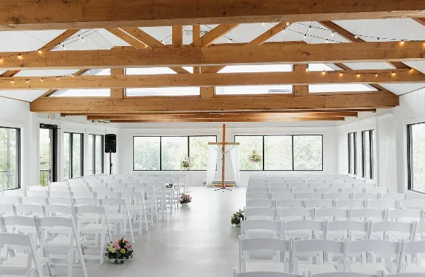 Interior view of a modern event center featuring exposed wooden ceiling beams, skylights, and rows of white chairs arranged for a wedding ceremony.