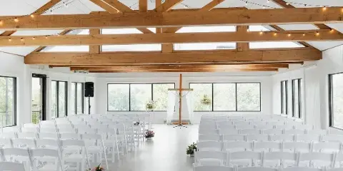 Interior view of a modern event center featuring exposed wooden ceiling beams, skylights, and rows of white chairs arranged for a wedding ceremony.