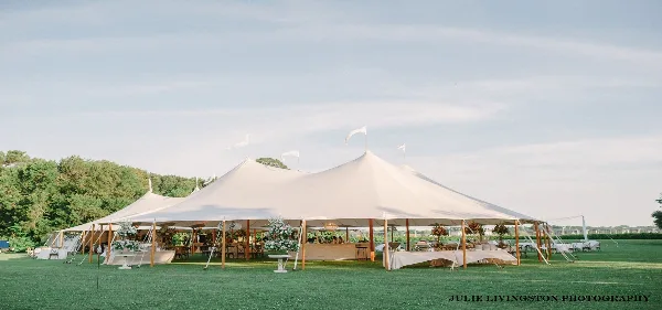 A large white event tent set up on a grassy field with tables, chairs, and floral centerpieces visible inside.