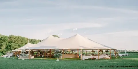 A large white event tent set up on a grassy field with tables, chairs, and floral centerpieces visible inside.