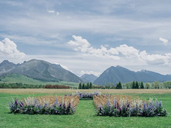 Outdoor wedding ceremony setup with rows of chairs and floral arrangements in a grassy field with mountains in the background.