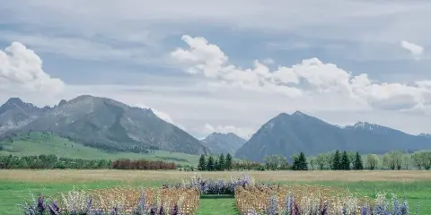 Outdoor wedding ceremony setup with rows of chairs and floral arrangements in a grassy field with mountains in the background.