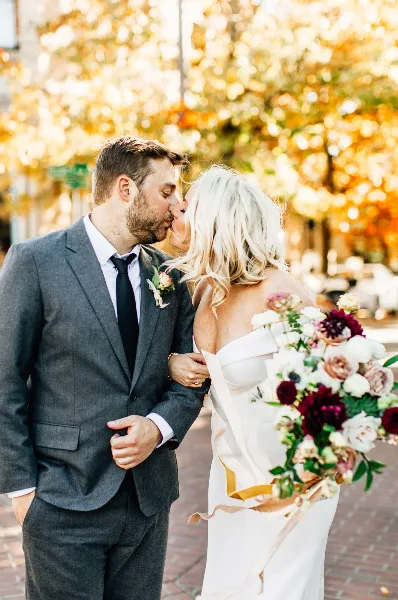 A bride and groom sharing a kiss outdoors with autumn foliage in the background.