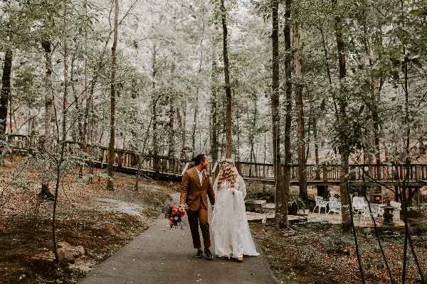A bride and groom walking on a paved path through a forest setting with a wooden bridge and white chairs in the background.