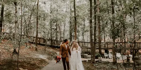 A bride and groom walking on a paved path through a forest setting with a wooden bridge and white chairs in the background.