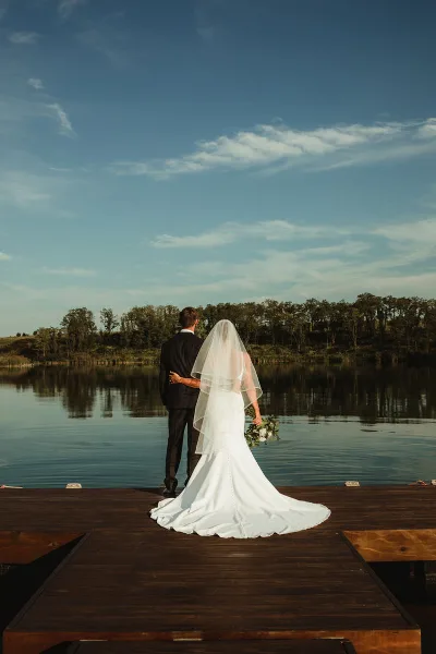 A bride and groom stand on a wooden dock overlooking a calm reservoir with a tree-lined shore in the background.