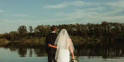 A bride and groom stand on a wooden dock overlooking a calm reservoir with a tree-lined shore in the background.