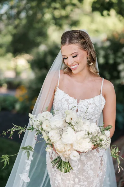 A smiling bride in a white lace wedding dress and veil, holding a bouquet of white flowers, with professionally styled hair and makeup.