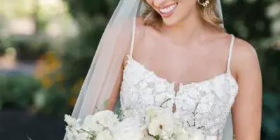 A smiling bride in a white lace wedding dress and veil, holding a bouquet of white flowers, with professionally styled hair and makeup.