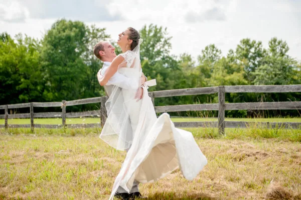 A bride and groom laughing and dancing together in a grassy field with a wooden fence in the background.