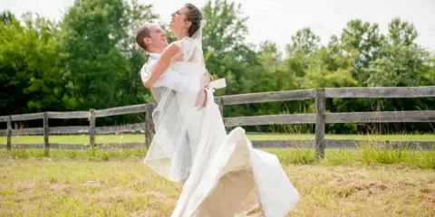 A bride and groom laughing and dancing together in a grassy field with a wooden fence in the background.