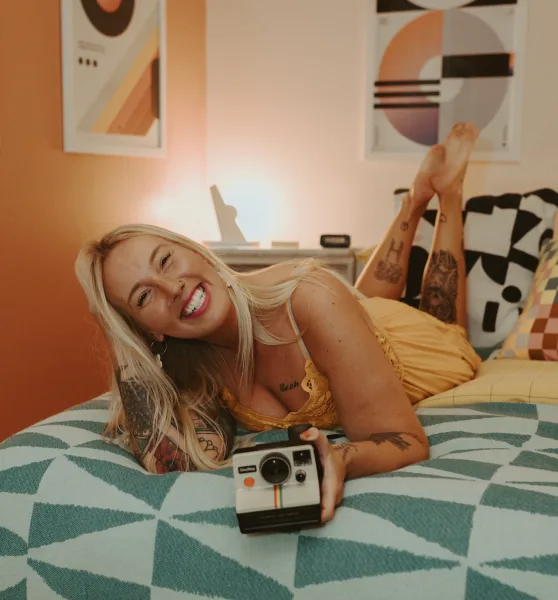 A smiling blonde woman with tattoos lies on a bed holding a vintage Polaroid camera.
