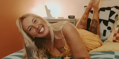 A smiling blonde woman with tattoos lies on a bed holding a vintage Polaroid camera.