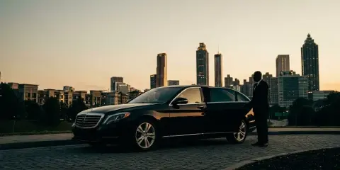 A black Mercedes-Benz executive sedan parked on a cobblestone street with a uniformed chauffeur standing by the open rear door, set against a city skyline at dusk.