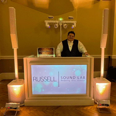 A DJ standing behind a professional booth with the Russell Sound Lab logo, flanked by tall speakers.