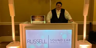 A DJ standing behind a professional booth with the Russell Sound Lab logo, flanked by tall speakers.