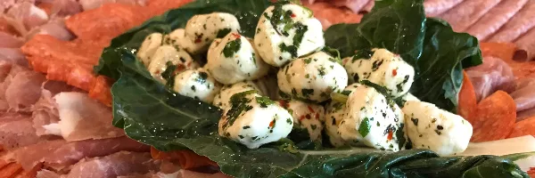 A catering platter featuring fresh mozzarella balls with herbs, served on a bed of dark green leaves surrounded by slices of cured meats.