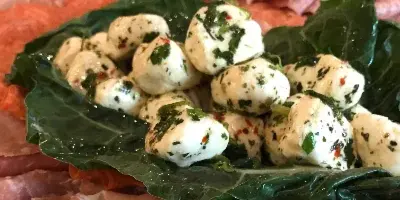 A catering platter featuring fresh mozzarella balls with herbs, served on a bed of dark green leaves surrounded by slices of cured meats.