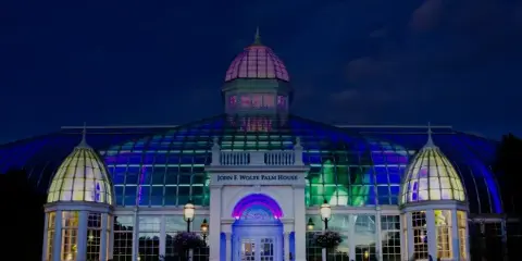 Nighttime exterior view of the John F. Wolfe Palm House at Franklin Park Conservatory, illuminated with colorful lights.