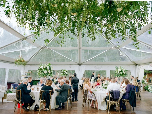 A wedding reception inside a clear-top tent featuring hanging floral installations and white table settings.