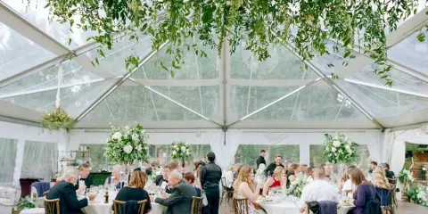 A wedding reception inside a clear-top tent featuring hanging floral installations and white table settings.