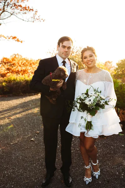 A wedding couple posing outdoors, with the bride holding a white and green floral bouquet.