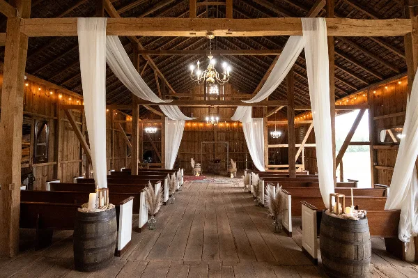 Interior of a rustic wooden barn wedding venue with pews, chandeliers, and white drapery.