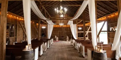 Interior of a rustic wooden barn wedding venue with pews, chandeliers, and white drapery.
