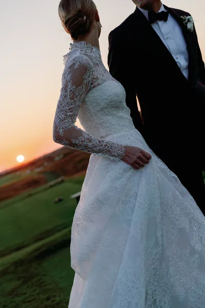 A bride in a lace wedding gown and a groom in a tuxedo standing together at sunset on a grassy hill.