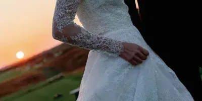 A bride in a lace wedding gown and a groom in a tuxedo standing together at sunset on a grassy hill.