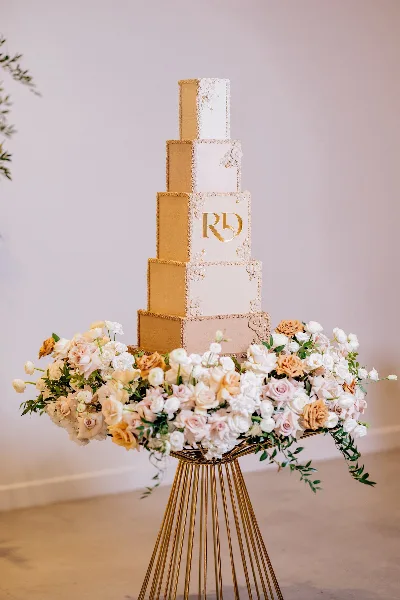 A tall, five-tiered wedding cake with gold and white finishes, featuring a gold monogram and surrounded by a large arrangement of white and peach roses on a gold stand.