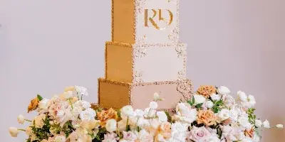 A tall, five-tiered wedding cake with gold and white finishes, featuring a gold monogram and surrounded by a large arrangement of white and peach roses on a gold stand.