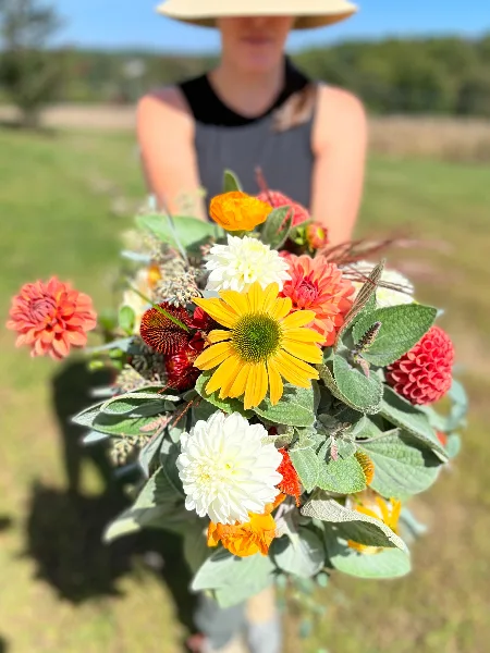 A person in a field holding a large bouquet of colorful seasonal flowers including daisies, dahlias, and sunflowers.