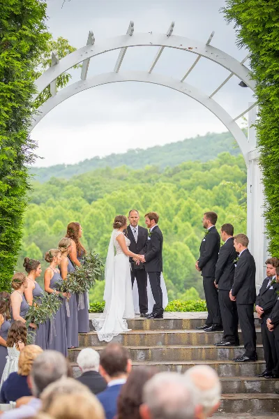 A bride and groom exchanging vows under a white arched trellis with a scenic mountain view in the background.
