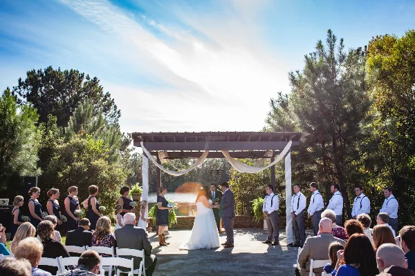 Outdoor wedding ceremony at a wooden pergola with a lake and pine trees in the background.