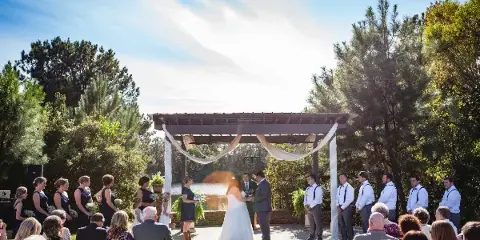 Outdoor wedding ceremony at a wooden pergola with a lake and pine trees in the background.