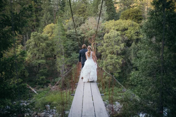 A bride and groom walking across a wooden suspension bridge in a forested area.