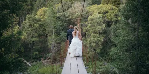 A bride and groom walking across a wooden suspension bridge in a forested area.