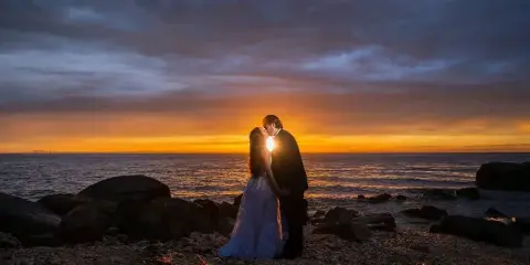 A bride and groom kissing on a rocky beach at sunset with the sun flaring between them.