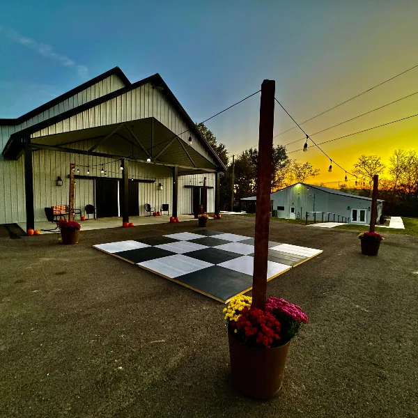Exterior view of a metal barn wedding venue with an outdoor dance floor and string lights at sunset.